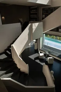 High-angle view of a modern open-concept living space featuring a sculptural white concrete staircase, dark floor, built-in bookshelves, and two white armchairs beside a large window overlooking a swimming pool.