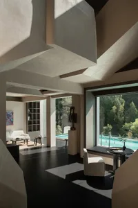 Modern minimalist living room featuring massive concrete architectural volumes, dark polished floors, white furniture, and large windows overlooking a swimming pool surrounded by forest.