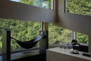 Modern concrete interior featuring a black hammock reading nook beside large windows with dense green forest view