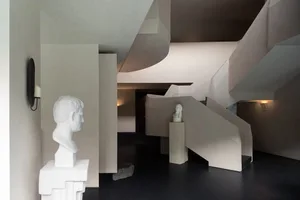 Modern interior featuring an angular concrete staircase, white classical marble busts on pedestals, and minimalist lighting.