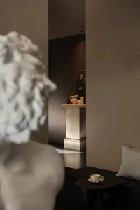 View of a contemporary minimalist living room featuring a travertine pedestal with decorative objects, a classical white marble bust in the foreground, dark flooring, and a built-in seating area with neutral cushions.