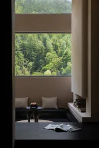 Modern minimalist reading nook with built-in window seat, neutral cushions, dark wood side table, and a large picture window overlooking a dense green forest.