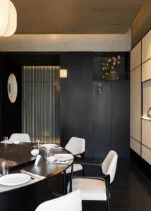 Modern dark-toned dining room featuring a large round black marble table, white upholstered dining chairs with dark wood frames, textured beige wall panels, vertical backlit slat wall feature, and pink cherry blossom arrangement in a built-in niche.