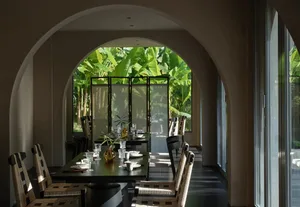Modern dining room featuring a large arched doorway that frames a tropical garden with banana trees, a dark dining table set with white ceramics, and chairs with black and cream woven rattan backs.