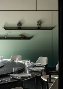 Modern dining room with round black marble table, white leather dining chairs, and floating shelves displaying decorative stones against a sage green gradient wall.