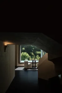 Modern minimalist living space featuring raw concrete architecture, a white armchair, round side table, and large window revealing a dense forest view.