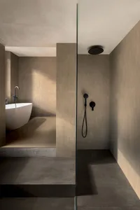 Modern minimalist bathroom featuring beige micro-cement walls and floors, a white freestanding bathtub on a raised platform, and a glass-enclosed walk-in shower with black matte fixtures.