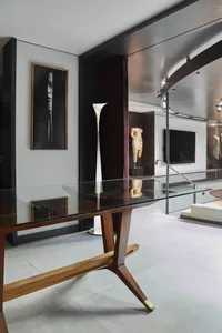 Modern living room interior featuring a walnut and glass console table, tall white floor lamp, framed black and white portrait, bronze classical torso sculptures, and a curved metallic ceiling detail.