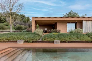 Modern terracotta villa with large glass openings, wooden elements, and a lush garden terrace beside a clear swimming pool under a blue sky.