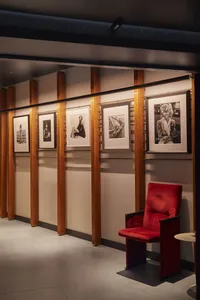 Modern gallery hallway featuring a vibrant red velvet armchair, walnut wood slat walls, and a series of framed black and white photographic portraits under warm track lighting.