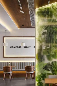 Modern cafe interior featuring walnut swivel chairs, grey upholstered booth seating, warm wood ceiling, and a glowing translucent green botanical partition wall.