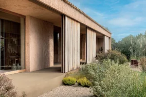 Side view of a modern rammed earth house featuring large glass windows, vertical timber sliding screens, and a gravel garden with native plants under a clear blue sky.