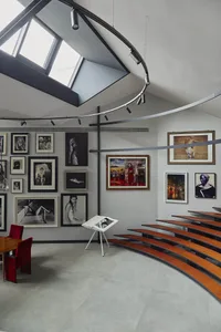 Modern art gallery interior featuring a curved multi-tiered oak amphitheater, large skylight, extensive framed photography collection on white walls, and red velvet seating area.