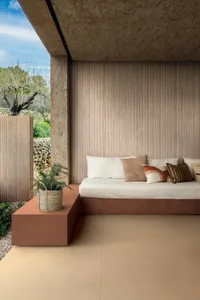 Modern outdoor lounge with a built-in terracotta daybed, white and earth-toned cushions, ribbed concrete wall, and potted olive tree under a clear blue sky.