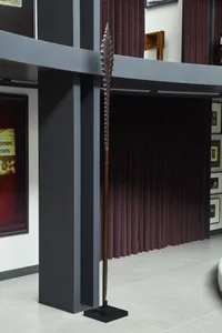 Modern living room interior featuring a tall tribal spear sculpture, dark grey structural beams, burgundy velvet curtains, and gallery-style framed art on white walls.