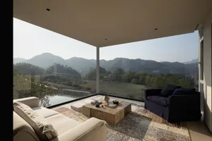 Modern living room with large floor-to-ceiling windows overlooking green mountains and a river, featuring a beige sofa, dark blue armchair, and wooden coffee table on a patterned area rug.