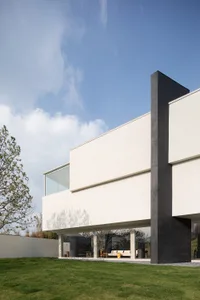 Modern minimalist concrete house featuring a large cantilevered white volume, dark grey structural elements, and extensive glass walls, set against a green lawn under a clear blue sky.