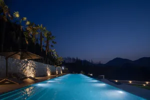 A luxurious infinity pool glowing with underwater lights at night, bordered by a textured natural stone wall, palm trees, and lounge chairs overlooking dark mountains.