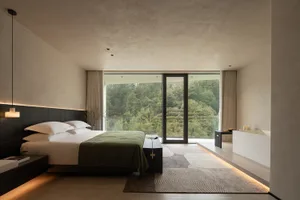 Modern minimalist bedroom featuring dark wood headboard, white bedding with green throw, concrete walls and ceiling, large floor-to-ceiling windows overlooking a dense forest, and a freestanding white bathtub on a raised platform.