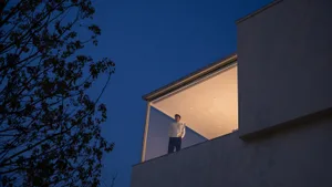 Man standing on a modern cantilevered concrete balcony at dusk with warm interior lighting against a deep blue sky and silhouetted tree branches.