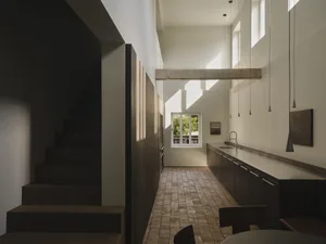 Modern minimalist kitchen and dining area with dark wood cabinetry, long white countertop, pendant lights, exposed concrete beam, and wooden staircase on the left.