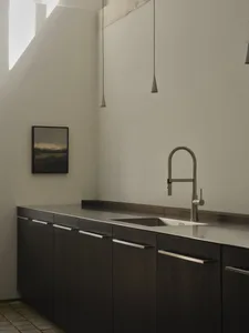 Modern minimalist kitchen featuring dark wood lower cabinets, stainless steel countertop and sink, high-arc faucet, concrete beam detail, and pendant lights with a framed landscape painting on the wall.