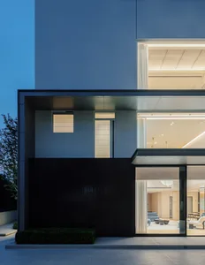 Exterior view of a contemporary luxury villa at dusk, showcasing dark wood paneling, large glass openings revealing an illuminated interior, and clean concrete architectural lines.