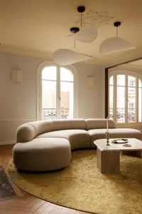A spacious Parisian living room featuring a large curved off-white boucle sectional sofa on a round chartreuse rug, with three sculptural white pendant lights hanging from a ceiling with ornate plasterwork, arched French windows, and herringbone wood flooring.