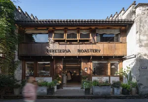 Street view of a two-story heritage building converted into 18 Reserva Roastery cafe, featuring dark reclaimed wood facade, large windows, open entrance, and numerous potted plants lining the stone pavement under a clear blue sky.