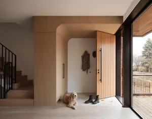 A light-colored curly-haired dog lies on the tiled floor of a modern minimalist entryway featuring light oak wood cabinetry, a curved architectural detail, black pivot front door, and large glass sliding doors opening to a wooden deck.