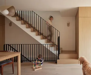 Modern family home interior featuring a light oak staircase with black metal railings, wooden dining table, pendant light, child playing, and a dog in the foreground.