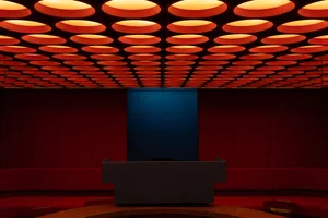 Modern theater auditorium featuring deep red velvet seating, a dark stage backdrop, and a ceiling covered in glowing orange circular light fixtures.