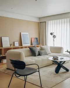 Modern minimalist living room with a large cream boucle L-shaped sectional sofa, black metal dining chair in foreground, round black coffee table, light wood wall paneling, and large window with sheer curtains.