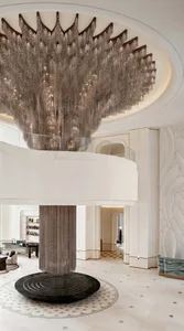 Expansive luxury hotel lobby featuring an enormous cascading crystal-bead chandelier hanging above a circular black marble base, with a curved mezzanine and light stone finishes.