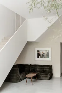 Modern minimalist living space featuring a dark green corduroy modular sofa under a white staircase, accompanied by a walnut coffee table and framed black and white photograph on the wall.