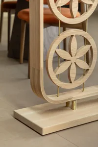 Close-up of a light oak room divider with circular floral cutout patterns, brass rods, and a curved wooden base, with orange dining chairs visible in the background.