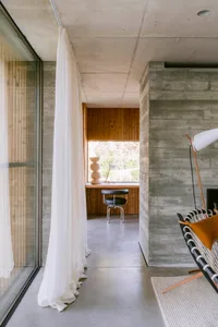 Modern concrete interior featuring a built-in wooden desk, black leather chair, sheer white curtain, and woven leather lounge chair in a sunlit reading nook.