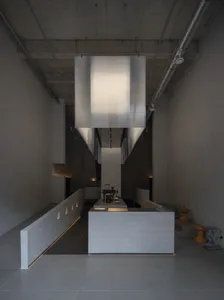 現代粗獷風廚房室內，裸露混凝土牆面、地板與天花板，大型矩形織物吊燈懸掛在混凝土中島上方，搭配外露金屬管線與整合式LED燈光。
