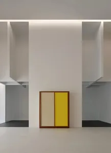 A framed two-tone panel with beige and bright yellow sections leaning against a white wall in a minimalist gallery space with dramatic architectural cutouts and hidden lighting.