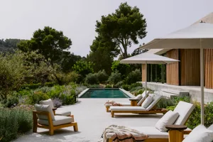 Modern minimalist poolside terrace featuring wooden lounge chairs with white cushions, large white umbrellas, concrete paving, and lush Mediterranean garden with olive trees and lavender under bright daylight.
