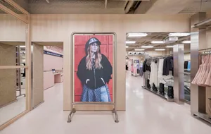 Modern fashion boutique interior featuring light wood paneling, a large digital portrait on a metal stand, stainless steel accents, and clothing racks in the background.