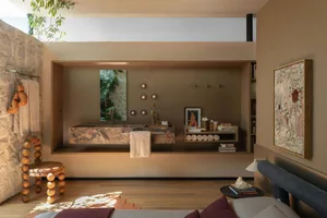 Modern earthy bathroom featuring a floating marble trough sink, stone wall with towel rack and boxing gloves, wooden bead stool, built-in shelving, and warm neutral tones.