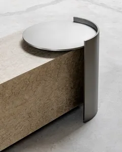 Close-up of a modern side table with a round brushed stainless steel top and a thick, curved base made of richly figured burl wood, resting on a polished concrete floor.