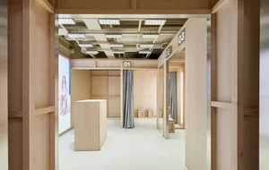 View into a modern minimalist retail fitting room area constructed with light-colored wood panels, featuring numbered changing rooms, grey curtains, a central wooden display block, and integrated LED lighting.