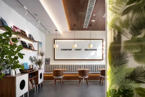 Modern cafe interior with wooden dining chairs, upholstered banquette seating, tropical leaf wallpaper, and a backlit sign reading COMFORT LIGHT DELICIOUS