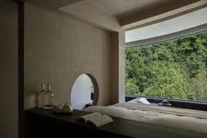 Modern minimalist bedroom interior with textured plaster walls, dark wood console, open book, and large window overlooking a green mountain forest.