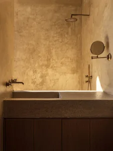 Modern minimalist bathroom featuring a concrete shower-bath, textured plaster walls, and warm brass fixtures with a round magnifying mirror.