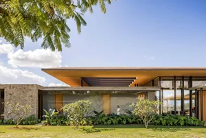 Modern tropical house exterior featuring a large cantilevered wooden roof, concrete walls, expansive glass windows, and lush tropical garden with flowering plants.