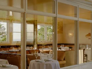Elegant seaside restaurant interior featuring marble walls, leather banquettes, round tables with white linens, and large windows overlooking the ocean.