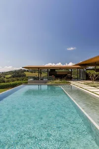Turquoise infinity pool with travertine deck extending toward a modern open-plan villa with large glass walls, set against a scenic rural landscape under a bright blue sky.